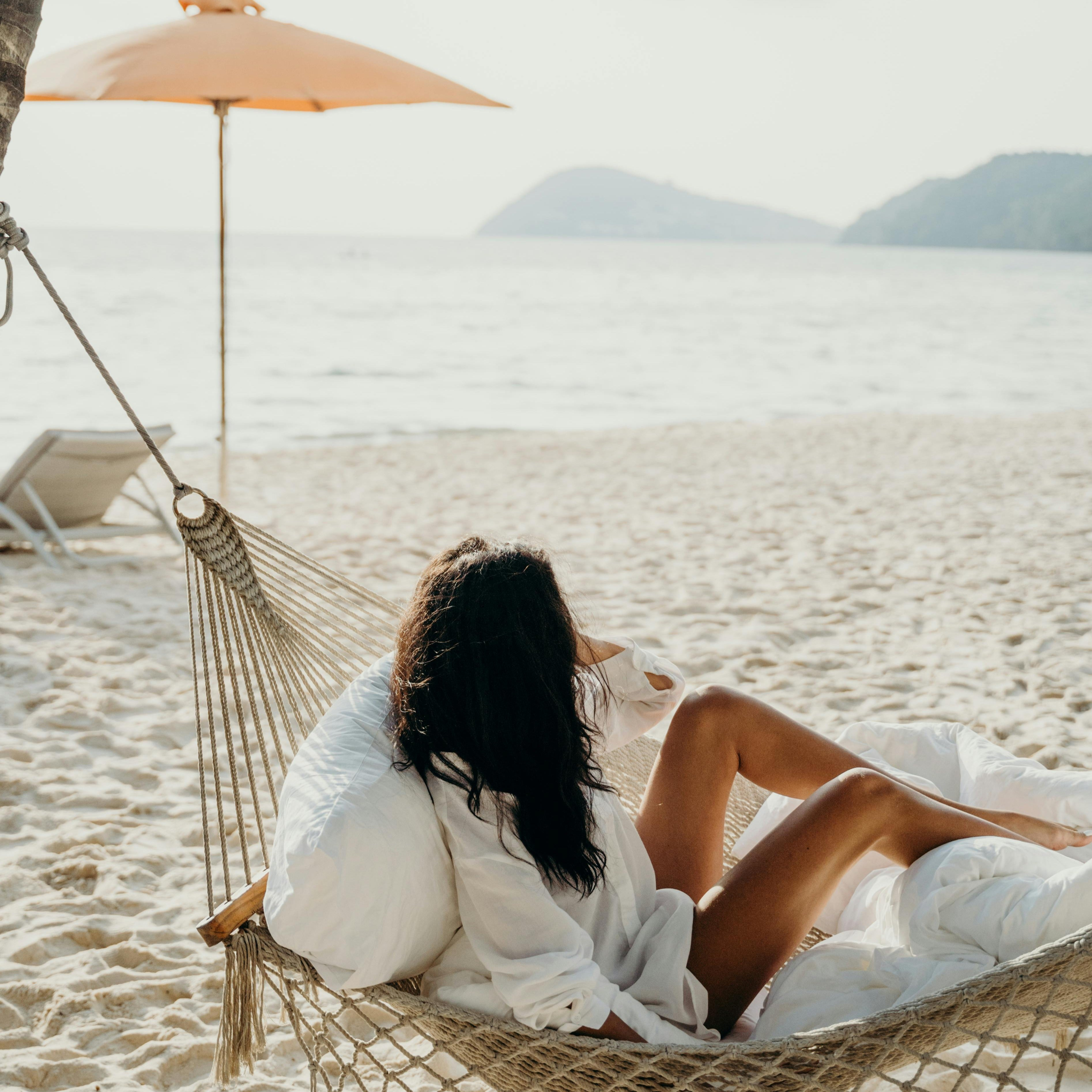 Woman relaxing in a hammock on the beach under a sun umbrella, ocean and mountains in the background.