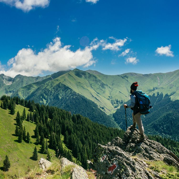 Hiker standing on a mountain peak with a backpack, surrounded by green valleys and blue sky, symbolizing outdoor adventure and exploration.
