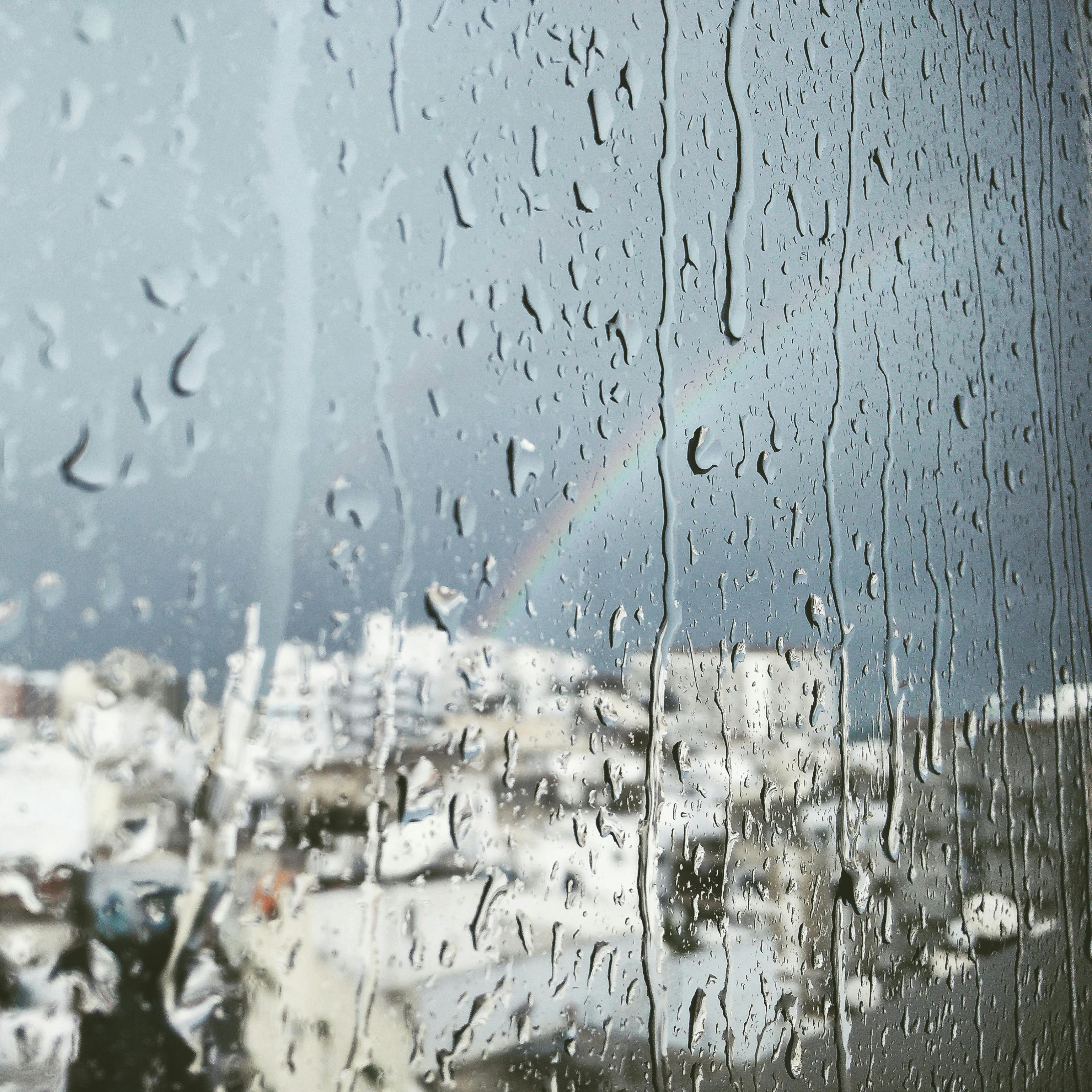 Rain drops on a window overlooking a city with a faint rainbow, symbolizing rainy weather and stylish rainwear.
