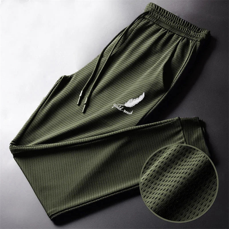 A pair of green ankle-length men's athletic pants with a drawstring waist, ankle ties, and a white logo on the left thigh. A close-up of the fabric shows a mesh-like texture.
