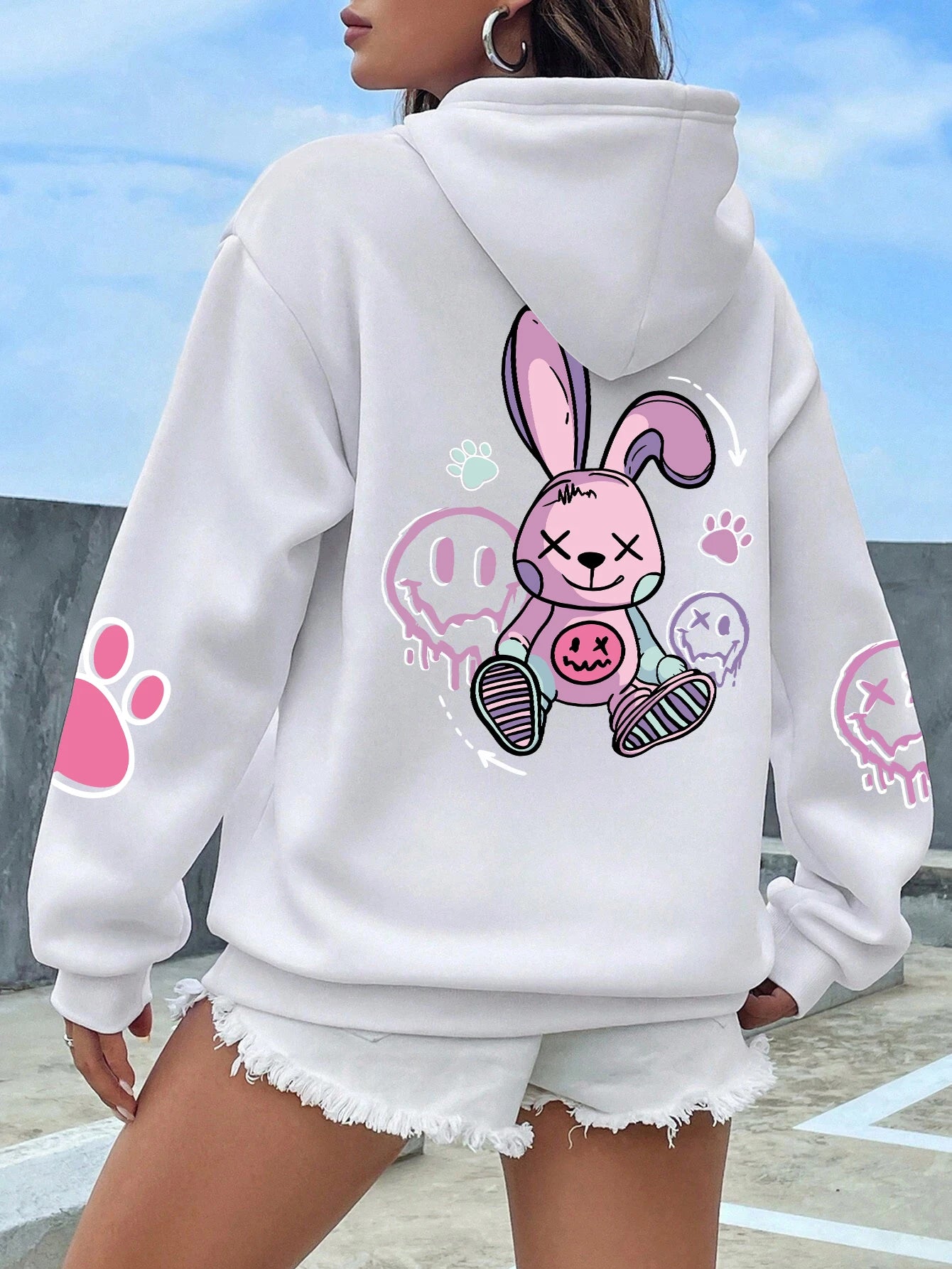 A female model wearing a white hooded sweatshirt with a colorful front print of a cartoonish rabbit character, paired with short denim shorts.