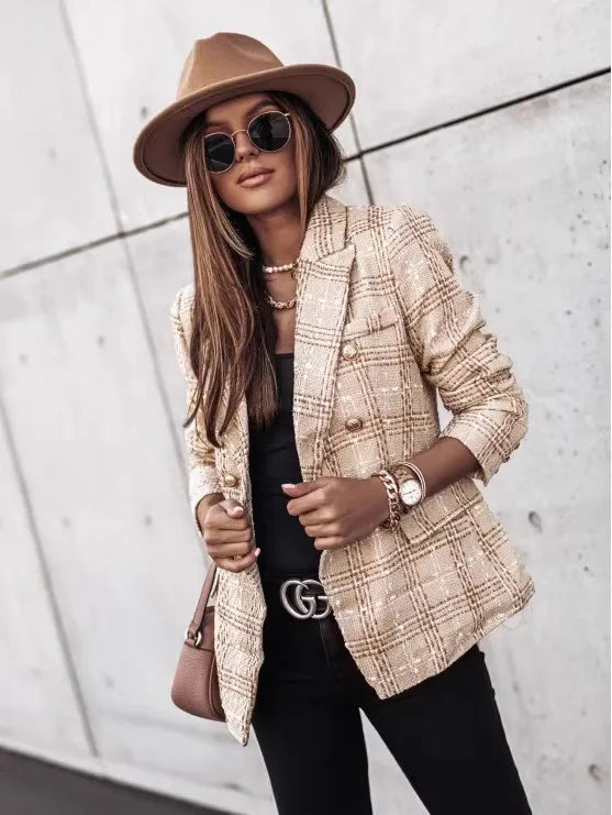Person wearing a beige plaid blazer, black sunglasses, and a brown hat against a light gray wall.