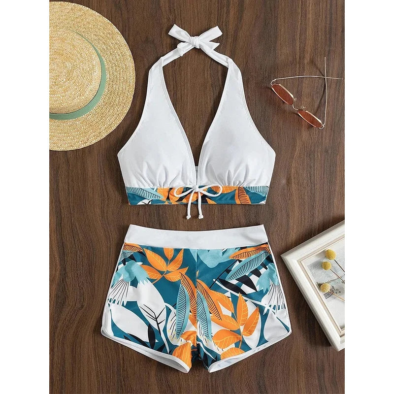 White bikini top and floral shorts on a wooden surface with a straw hat and sunglasses.