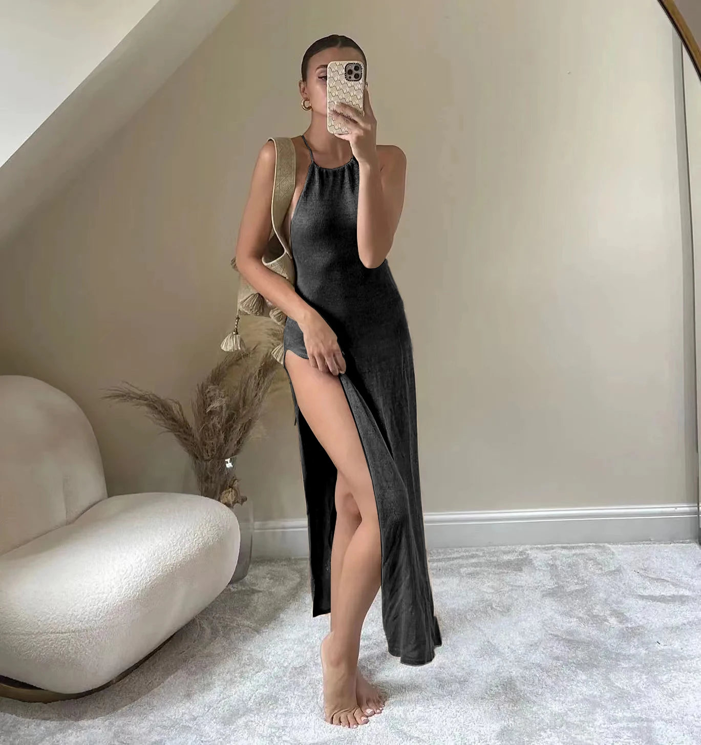 Woman in a black slip dress taking a mirror selfie in a room with a white sofa and decorative plant.